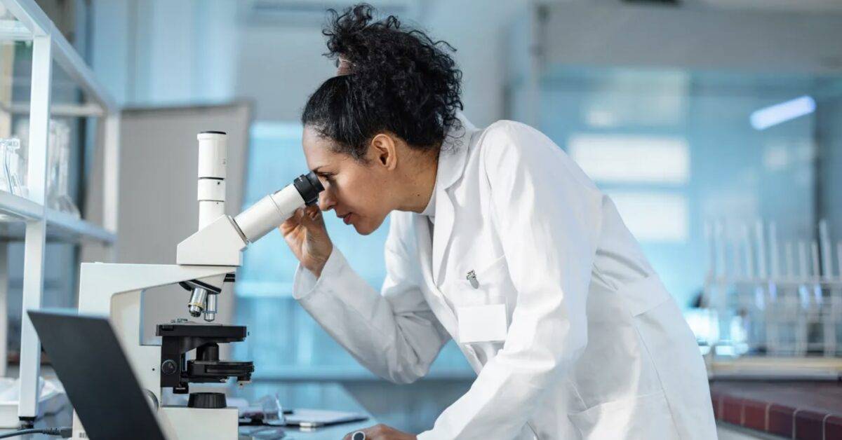 Female Scientist Looking Under Microscope And Using Laptop In A Laboratory