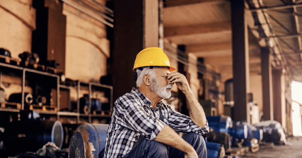 A worried senior factory worker is sitting next to the machines and holding his head. He is hoping the work he has done is fine in the asbestos plant